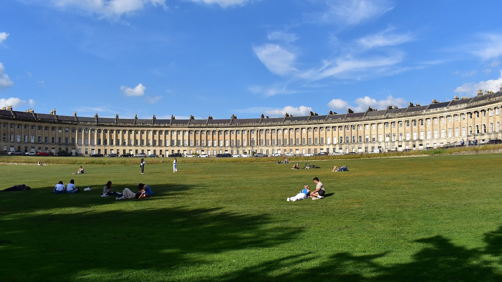 The Royal Crescent, Bath - iconic curved Georgian terrace seen across its green lawn, bathed in sunlight with people relaxing