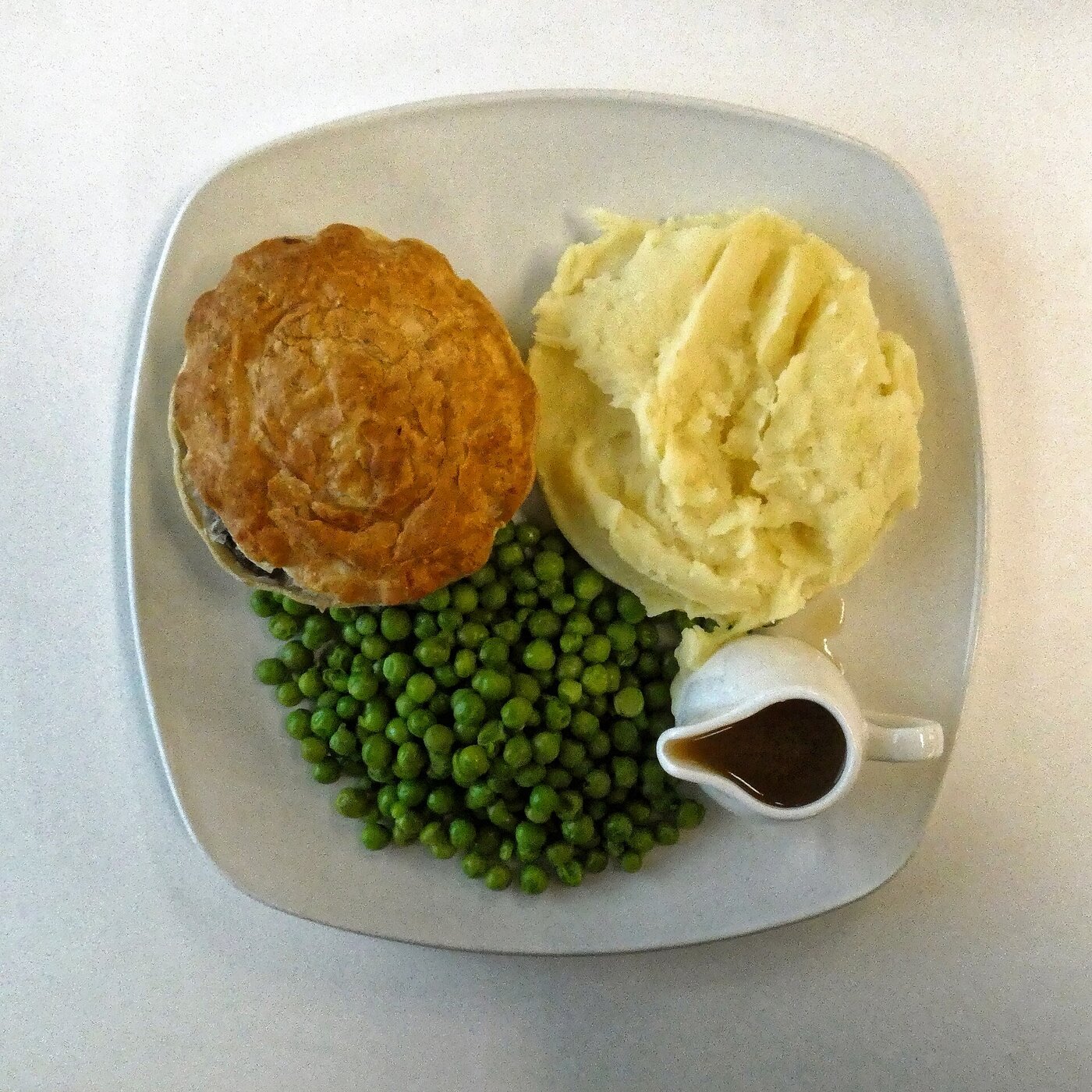 A plated British pub classic: a homemade pie with mashed potato, garden peas and a jug of gravy