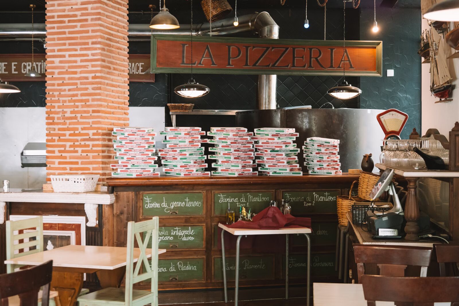 A rustic Italian pizzeria interior with a wood counter, wood-fired pizza oven, and stacked pizza boxes